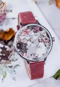 Montre ronde avec un cadran à motif floral, boîtier en argent et bracelet en cuir rose. Comprend des marqueurs en cristal et de fines aiguilles argentées.