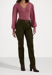 Růžový žebrovaný svetr s vycpanými rukávy, kombinovaný se tmavě zelenými cargo kalhotami s postranními kapsami a hnědým páskem. Hnědé kotníkové boty na podpatku.