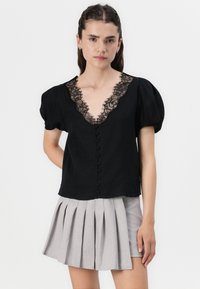 Top nero con maniche a palloncino e profondo scollo a V bordato in pizzo, arricchito da bottoni decorativi. Abbinato a una gonna plissettata grigia chiara.
