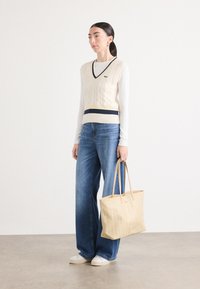 Beige Kabelstrick-Westen mit navyblauen und gelben Streifen, kombiniert mit lockeren blauen Jeans und einer beigen Tragetasche mit Muster.