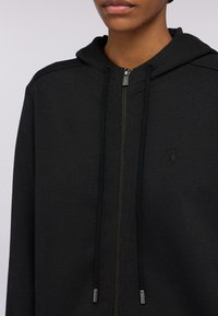 Černá zipová mikina vyrobená z měkké tkaniny. Obsahuje kapuci na stahování, zipové zapínání a embosované logo na hrudi. Hladká textura.