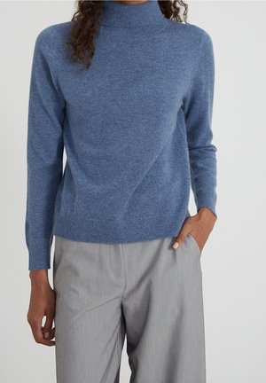Personne portant un pull à col roulé bleu et un pantalon gris clair, avec une main dans la poche et les cheveux bouclés partiellement visibles.
