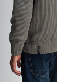 Grijze geribde trui met gestructureerde stof en een aansluitende zoom. Een zwart etiket met witte tekst is zichtbaar bij de onderkant. Donkere spijkerbroek met gestikte zakken.