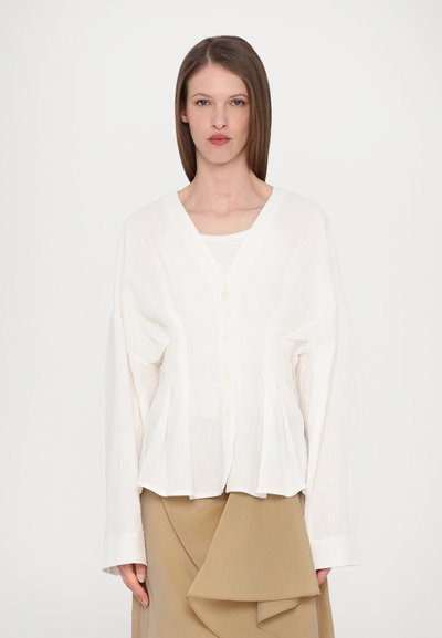 White, long-sleeve blouse with a v-neckline, featuring a button front, pleat details, and a relaxed fit, paired with a beige skirt.