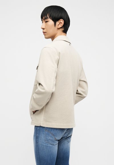 Νεαρός άνδρας με κοντά μαύρα μαλλιά, που φοράει μπεζ μπουφάν και μπλε τζιν, στέκεται με τα χέρια στις τσέπες μπροστά σε απλό λευκό φόντο.