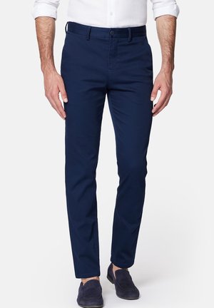 Mann trägt dunkelblaue Slim-Fit-Hose, weißes Hemd mit hochgekrempelten Ärmeln und dunkelblaue Wildleder-Loafer, steht vor weißem Hintergrund.