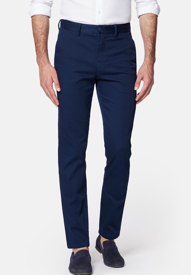 Homme portant un pantalon bleu marine slim, une chemise blanche aux manches retroussées et des mocassins en daim bleu foncé, debout sur un fond blanc.