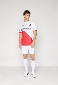 Mann, der in weißem und rotem Fußballtrikot mit weißen Socken und Stollenschuhen vor einem schlichten grauen Hintergrund steht.