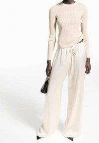 Donna che indossa un top a coste beige con maniche lunghe, pantaloni larghi crema a vita alta, scarpe nere a punta e tiene in mano una piccola borsa nera.