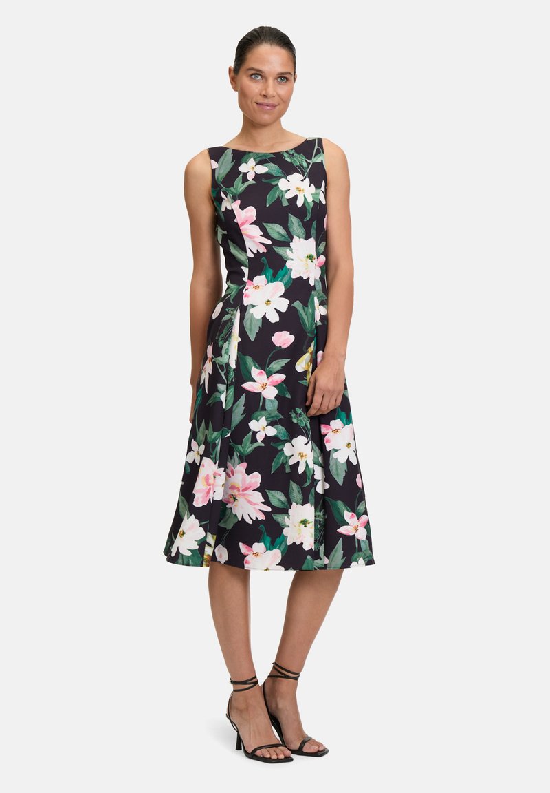 Robe fleurie avec un fond noir ornée de fleurs roses et blanches, sans manches, silhouette en A, et longueur mi-mollet. Accessoirisée avec des escarpins noirs.