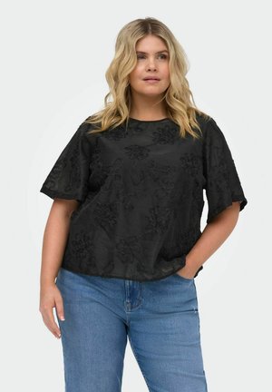 Frau mit welligem blondem Haar, die ein schwarzes, strukturiertes, geblümtes Oberteil und blaue Jeans trägt, steht mit einer Hand in der Tasche vor einem neutralen Hintergrund.