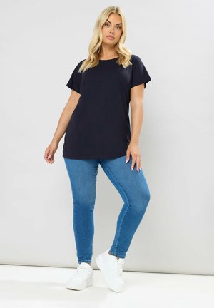 Femme blonde debout contre un mur uni, portant un haut noir à manches courtes, un jean skinny bleu et des baskets blanches.
