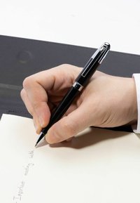 Juodas rašiklis laikomas rankoje, rašantis ant kreminio popieriaus. Fone tamsi tekstūruota paviršius. Rankoje matomas lengvas sukibimas ir natūralus odos tonas.