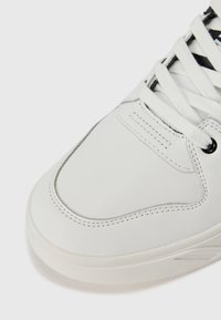 Sneaker atlético de couro branco com superfície lisa, biqueira arredondada, detalhes de costura sutis e atacadores planos. Sola de borracha flexível.