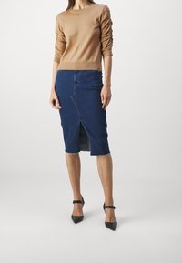 Mujer vestida con una blusa de manga arruchada color beige, una falda lápiz de mezclilla azul con abertura frontal y tacones de punta negra con tira en el tobillo, de pie sobre un fondo blanco.