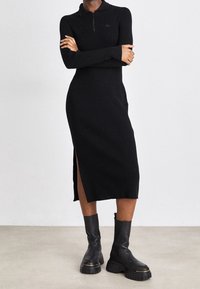 Robe en tricot côtelé noir avec un col zippé et des fentes latérales. Associée à des bottes noires à semelle épaisse. Détail de marque minimaliste sur la poitrine.
