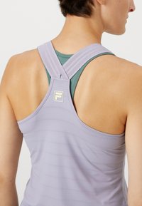 Fialová športová tank topka so štruktúrovaným vzorom. Má krížové ramienka a malé logo na chrbte. Obliekaná cez tyrkysovú blúzku.