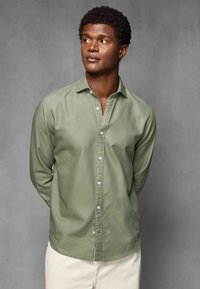 Grünes Langarmhemd aus leichtem Stoff mit subtiler Textur. Mit einem button-down Kragen und weißen Knöpfen. Schlichtes Design.