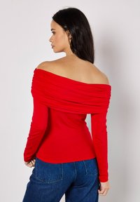 Rotes Oberteil mit schulterfreiem Schnitt, geraffter Textur, langen Ärmeln und taillierter Passform. Kombiniert mit blauen Jeans.
