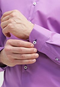 Fioletowa koszula z gładką teksturą, z białymi guzikami i kontrastującymi granatowymi akcentami na mankietach. Ręka poprawiająca mankiet.