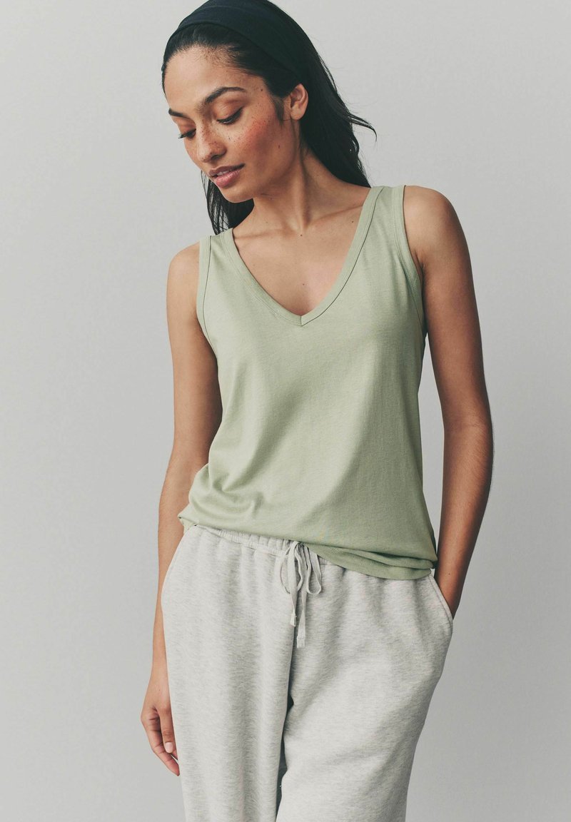 Top sin mangas de color verde claro con escote en V, hecho de tela suave, combinado con pantalones de chándal grises que tienen una cinturilla elástica y bolsillos laterales.