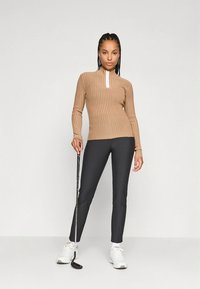 Beige ribbad långärmad tröja med en vit kvartszipp, tillsammans med smala svarta byxor och vita träningsskor, håller i en golfklubba.