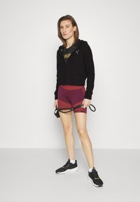 Felpa corta nera con zip sopra una maglietta scura, abbinata a pantaloni corti testurizzati color marrone scuro e scarpe nere. Il modello tiene una tracolla nera.