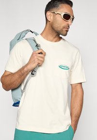 Εκρού κοντομάνικη μπλούζα με τσέπη στο αριστερό στήθος, με πράσινο λογότυπο "Patagonia". Η εμφάνιση ολοκληρώνεται με ένα ανοιχτό μπλε σακίδιο πλάτης και γυαλιά ηλίου.