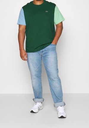 T-shirt à manches courtes bloc de couleur verte et bleue avec le logo Lacoste, associé à un jean bleu délavé et des baskets blanches.