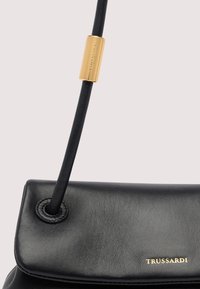 Bolso de cuero negro con acento cilíndrico dorado en la correa, grabado con "TRUSSARDI" y logo impreso en la solapa frontal.
