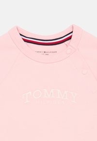 Hanorac roz deschis cu logo brodat "TOMMY HILFIGER", având un guler striat cu dungi roșii și bleumarin și închidere cu capse.