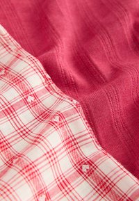 Gros plan sur deux tissus, l'un rouge avec des nervures texturées et l'autre blanc à motif écossais rouge avec de petits détails brodés.