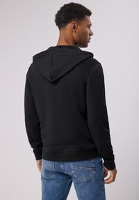 Černá mikina s kapucí, měkká na dotek, s žebrovanými manžetami a rovním lemem, oblečená s světle modrými džíny. Viditelná detail kapuce.