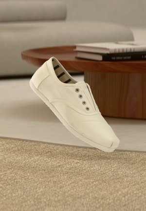 Witte canvas sneaker met vijf metalen veterogen, gestreepte binnenvoering, afgeronde neus en een gestructureerde rubberen zool. Opgehangen boven een geweven oppervlakte.