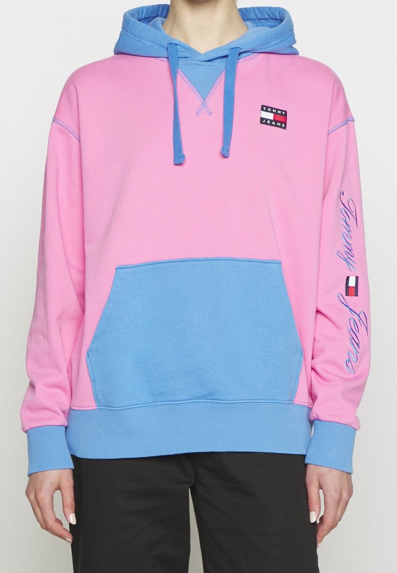 Personne portant un sweat à capuche Tommy Jeans rose et bleu avec poche avant, capuche avec cordons, et logo sur la poitrine et la manche droite.