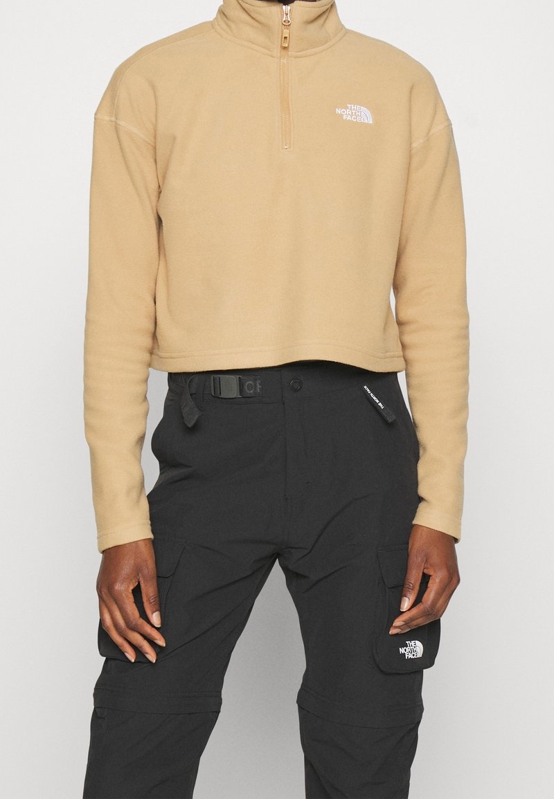 Pull polaire court beige avec zip quart de fermeture et pantalon cargo noir avec ceinture et poches à rabat, les deux avec les logos The North Face.