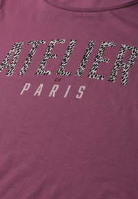 T-shirt violet avec le texte "ATELIER DE PARIS", les lettres "ATELIER" ont un motif léopard noir et blanc.