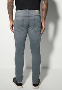 Jeans din denim gri, cu croială slim-fit, dotate cu două buzunare la spate și detalii de cusătură. Modelul poartă adidași negri, evidențiind o textură netedă a materialului.