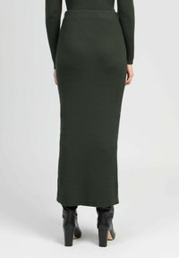 Jupe maxi en tricot côtelé vert foncé avec une taille ajustée, des fentes latérales, associée à des bottes à talons hauts noires. Texture lisse.