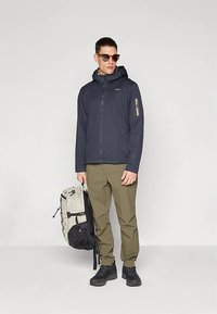 Veste légère bleu marine avec capuche, poche zippée, pantalon beige, chaussures noires et sac à dos gris-noir. Le modèle porte des lunettes de soleil.