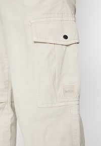 Béžové kapsáčové kalhoty s kapsou na stehně, černými kovovými doplňky a čtvercovou štítkem s nápisem "BOSS" v šedé výšivce.