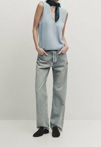 Vaaleansininen hihaton paita, jossa on V-pääntie, yhdistettynä löysiin, vaaleanharmaisiin farkkuihin ja tummanruskeisiin kenkiin. Musta huivi solmittuna kaulan ympäri.