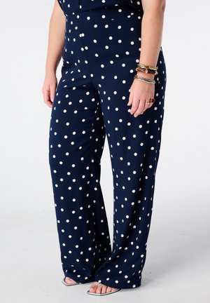 Vrouw draagt marineblauwe wijde broek met witte stippen, zilveren sandalen met open teen en armbanden aan haar rechterpols.