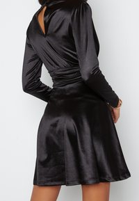 Robe en satin noire avec des manches longues, taille cintrée et jupe évasée. Présente un dos en trou de serrure et une texture lisse et brillante.