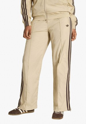 Pantaloni da tuta beige con strisce laterali marroni e logo Adidas, indossati con giacca da tuta abbinata e sneakers marroni con strisce bianche.