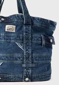 Dunkelblaue Denim-Tasche mit hellen Nähten, ausgestattet mit mehreren Taschen, einer strukturierten Oberfläche und einem auffälligen Markenetikett an der Oberseite.