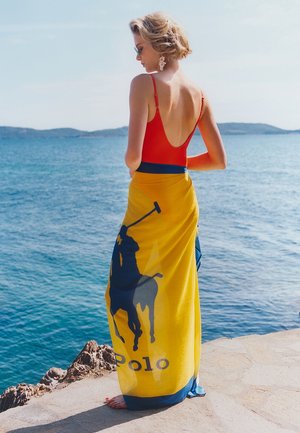 Femme aux cheveux blonds courts portant un maillot de bain rouge et un châle jaune Polo Ralph Lauren, debout pieds nus au bord de l'océan par une journée ensoleillée.