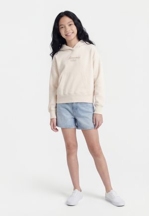 Smilende pige stående, iført beige Abercrombie-hættetrøje, lyseblå denimshorts og hvide sneakers mod en hvid baggrund.