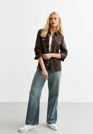 Veste en chemise marron avec fermeture à boutons, deux poches poitrine, associée à un jean bleu clair à jambes larges et des baskets argentées. Texture de tissu lisse.