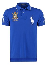 Blå pikétröja i bomull med texturerad yta. Har en vit broderad logotyp, kontrasterande krage och dekorativ sömnad.